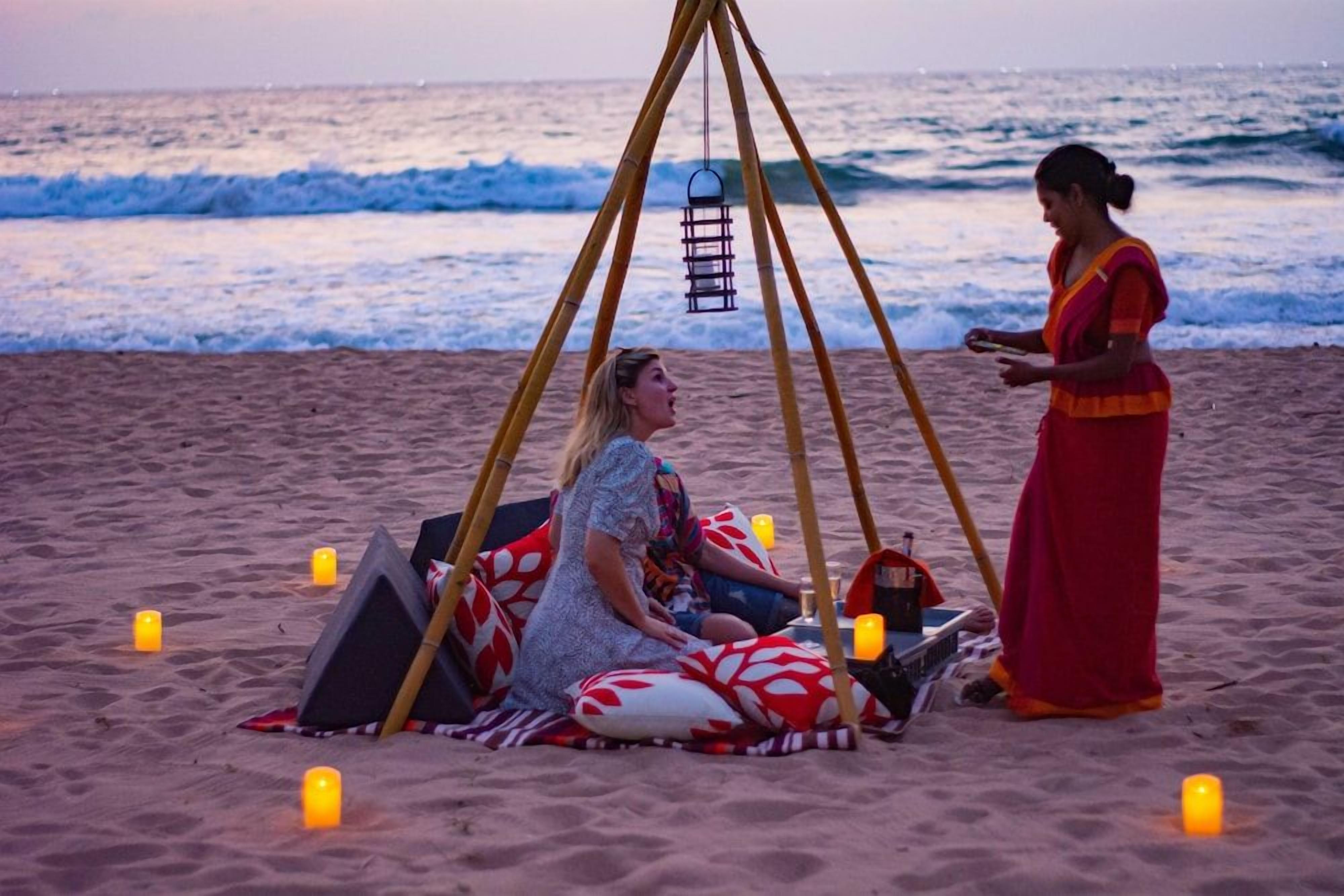 Une scène de plage avec une tente et des bougies, une personne assise à l’intérieur et une autre debout à l’extérieur.