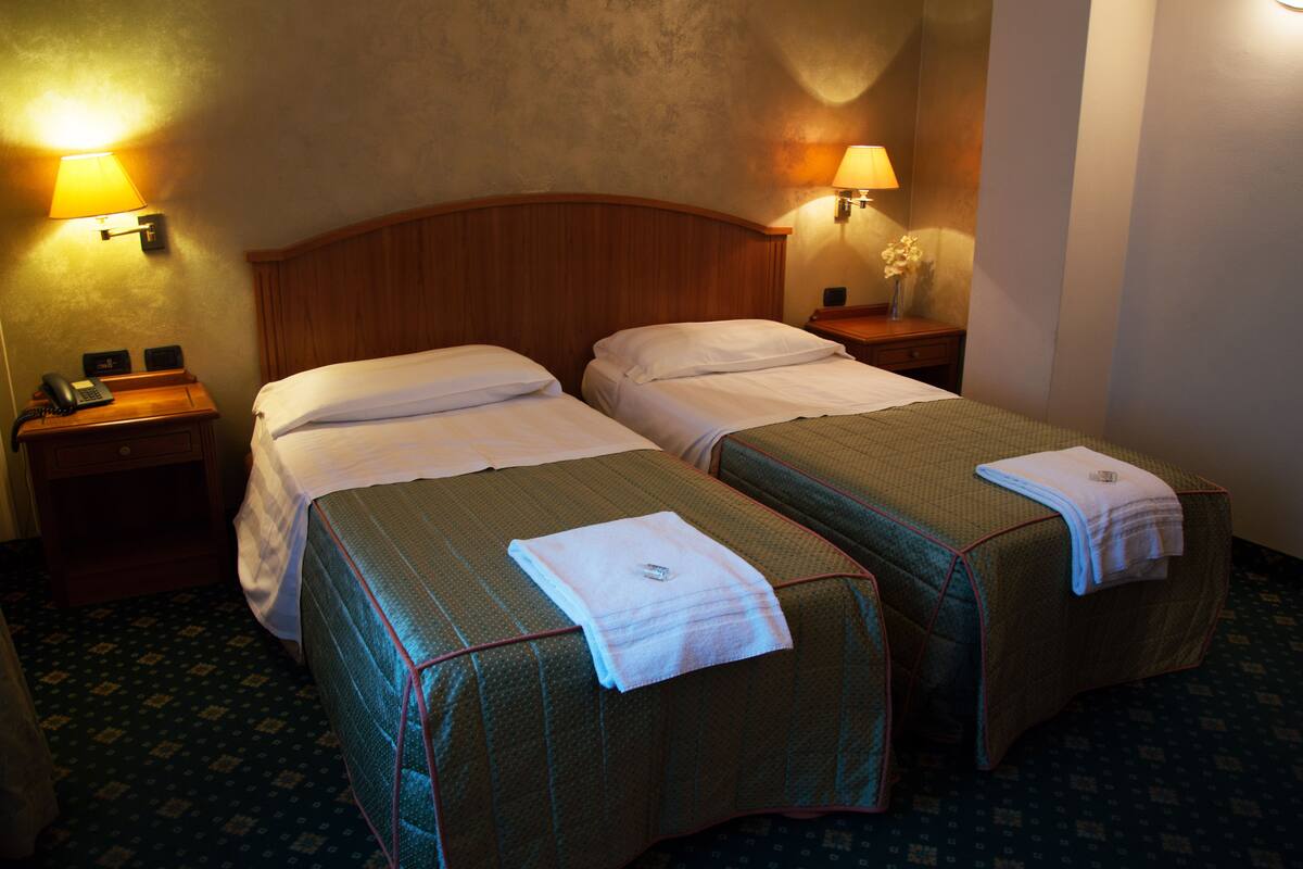 Une chambre d’hôtel avec deux lits simples, chacun muni d’un oreiller et d’une serviette pliée posée sur le lit. Il y a deux tables de chevet, l’une avec une lampe et l’autre avec un téléphone et une petite table. La chambre est dotée d’une tête de lit en bois et d’un tapis à motifs.