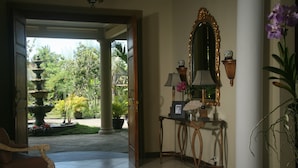 A room with a chair, a table with a lamp, a mirror, and a book. There is a door leading to an outdoor area with plants and a fountain. - Villa Pakem (Pakem)