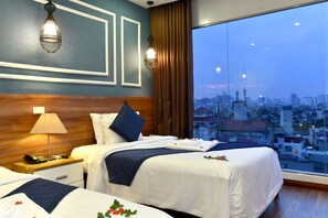 A hotel room with two beds, a wooden headboard, a nightstand with a lamp, and a city view through large windows. - Eternity Hanoi Hotel (Hanoi)