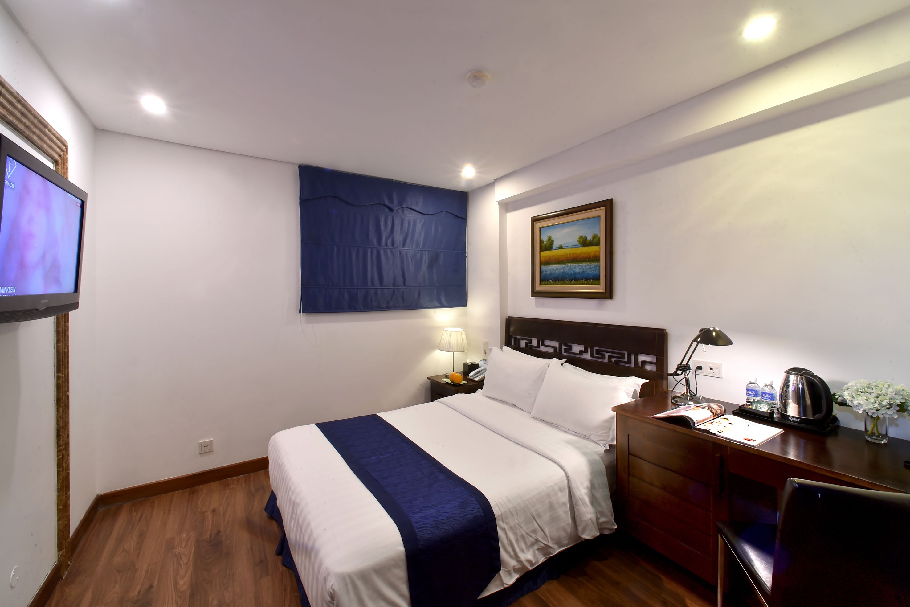 A hotel room with a bed, a desk, a chair, a nightstand, a lamp, a painting, and a television.