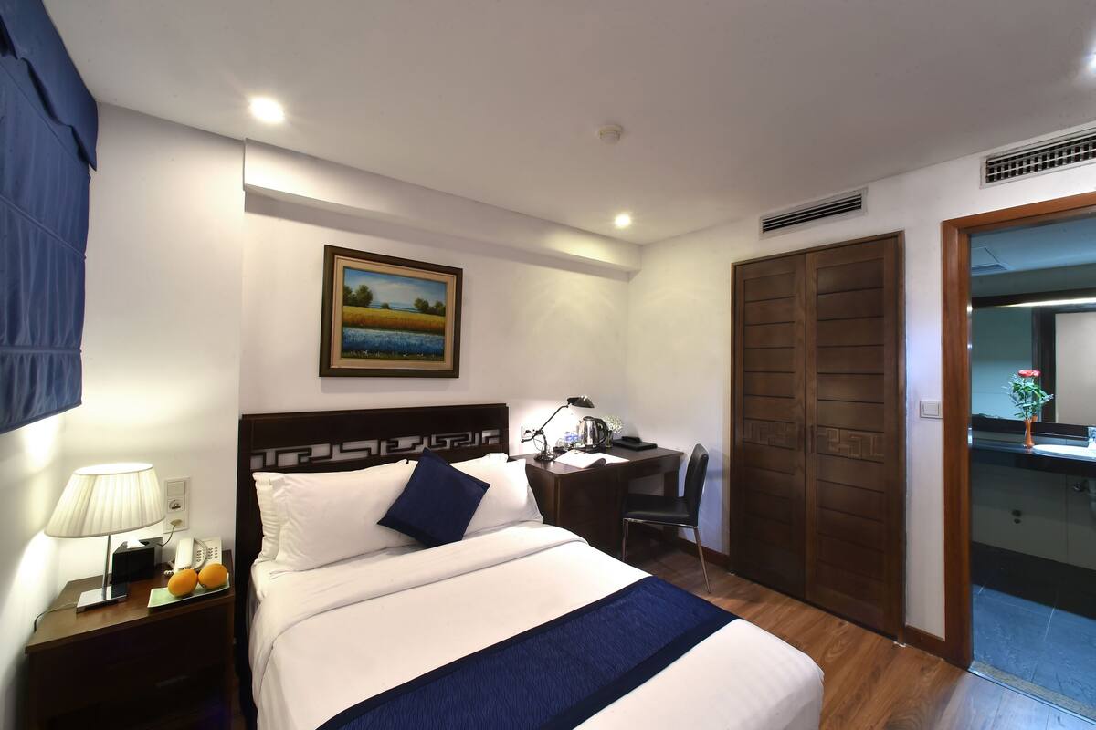 A hotel room with a bed, a desk, a chair, a lamp, and a painting on the wall.