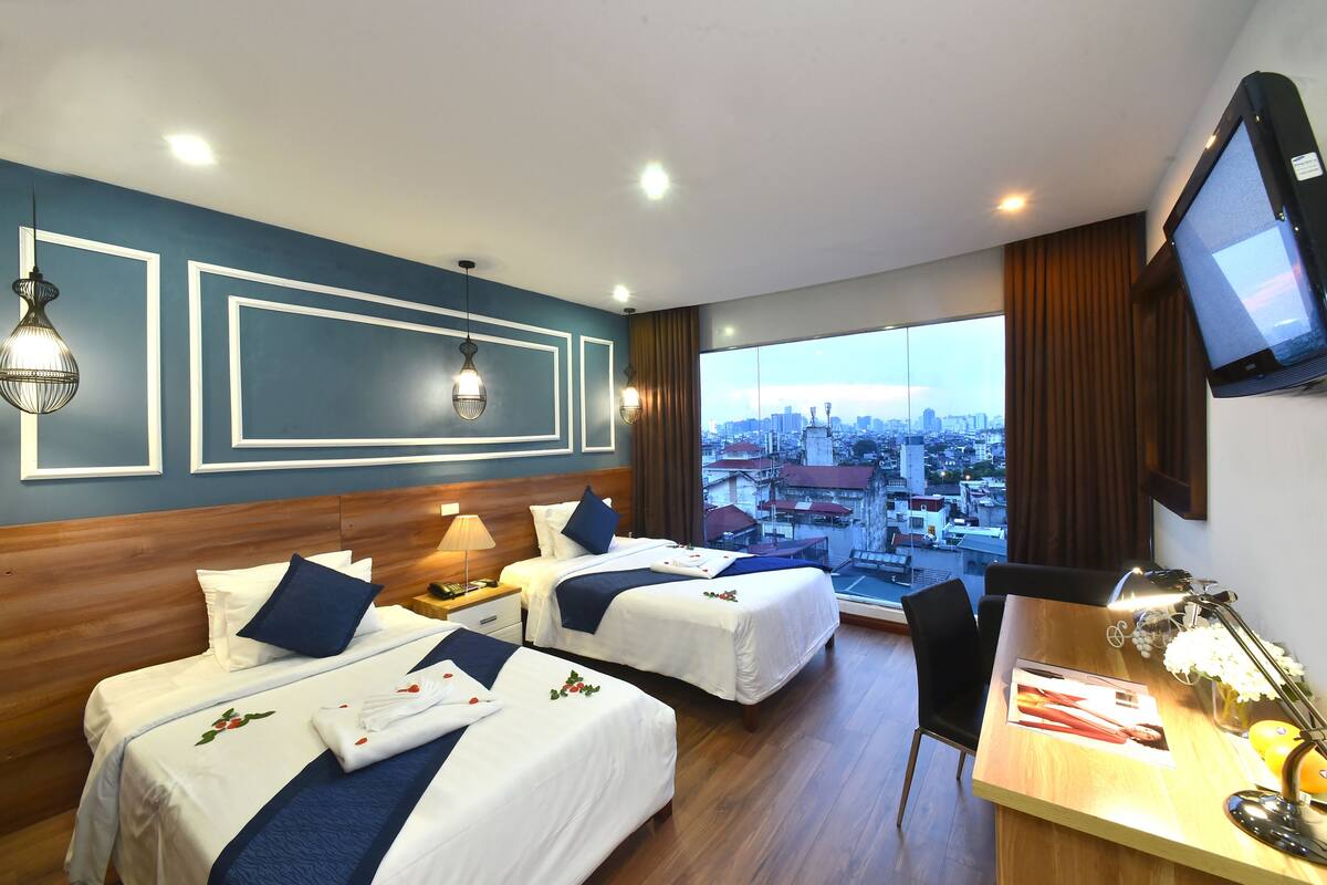 A hotel room with two beds, a desk with a lamp, a TV, and a large window with a city view.