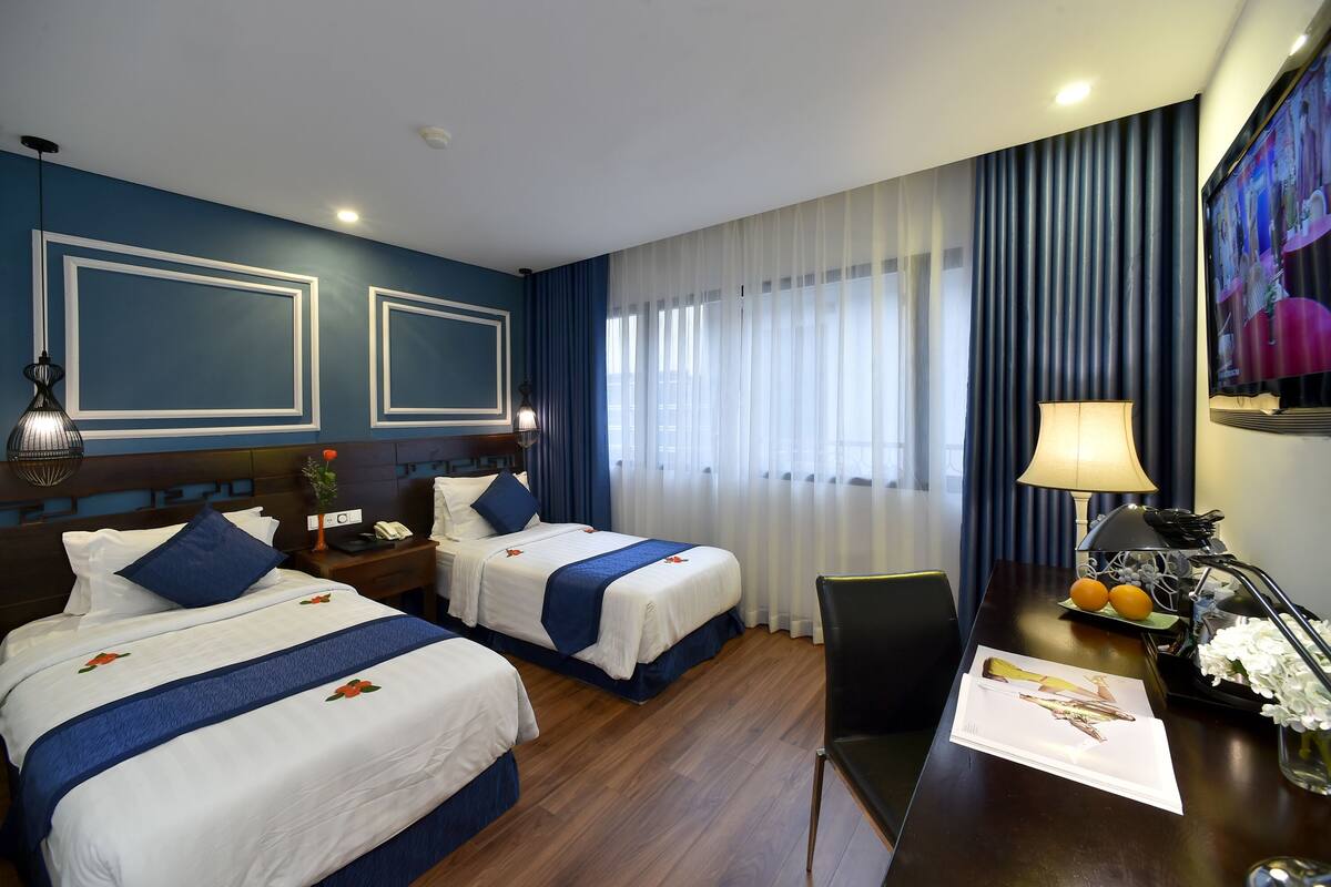 A hotel room with two beds, a desk with a lamp, a TV, and a window with curtains.