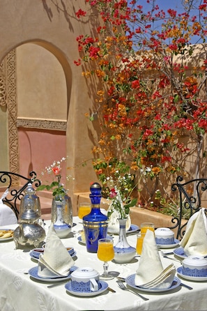 A table set for a meal with blue and white tableware, silver and blue vases, and a backdrop of a wall with flowering plants.