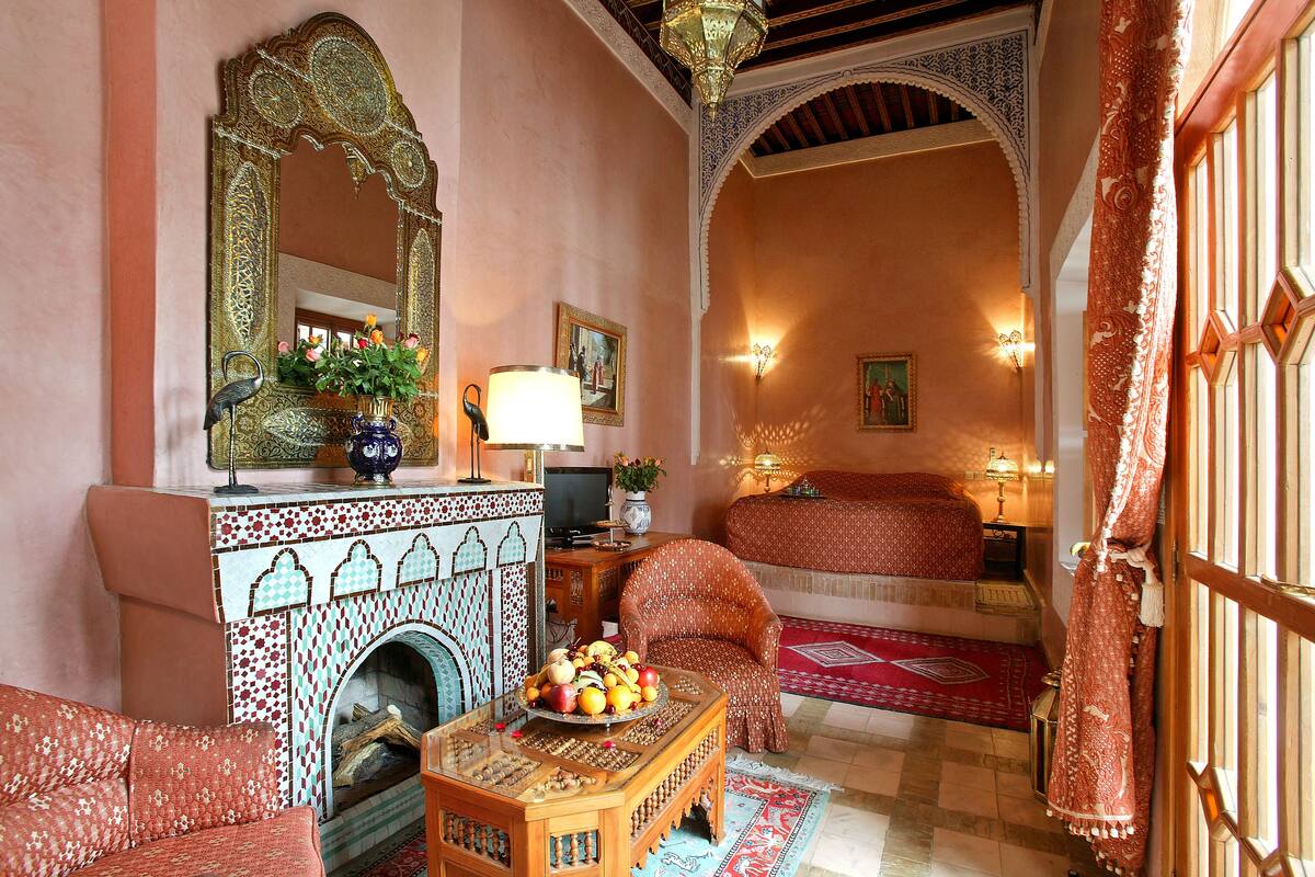 A traditional Moroccan room with a sofa, a table with fruit, a fireplace, and a bed with a red and white patterned bedspread.