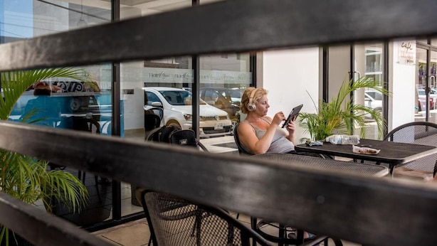 Una persona sentada a una mesa con una computadora portátil, frente a una ventana que refleja coches estacionados y un edificio con el rótulo 'Hotel Pizza'.