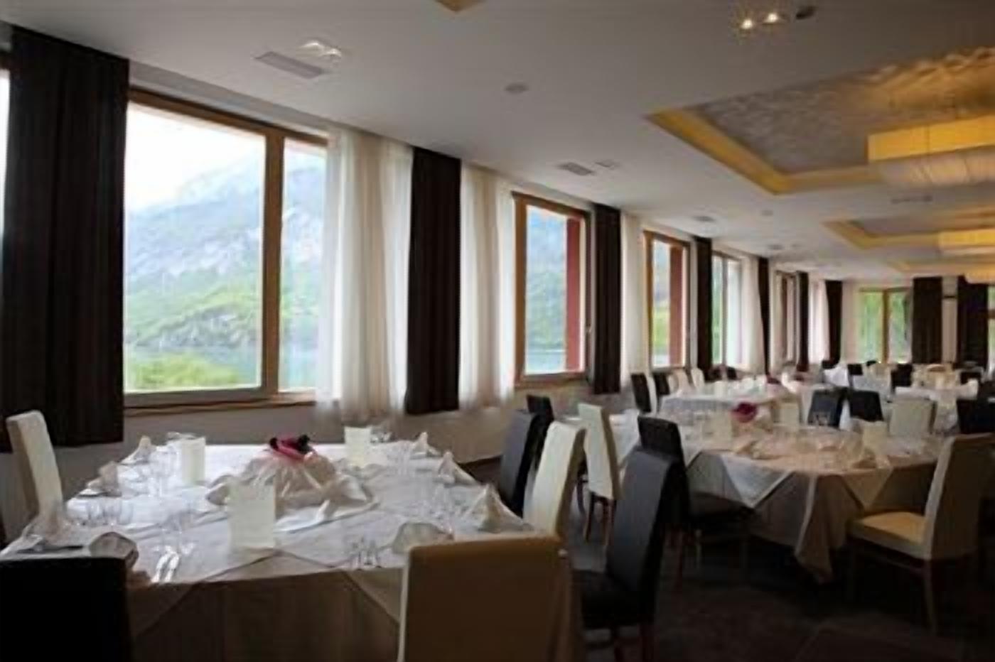 A dining area with tables set for a meal, large windows with curtains, and a view of the outdoors.
