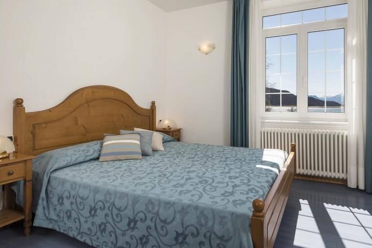 A bedroom with a wooden headboard, a patterned bedspread, a nightstand with a lamp, and a view of mountains through a window with curtains.