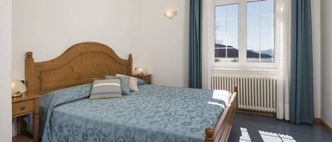 A bedroom with a wooden headboard, a patterned bedspread, a nightstand with a lamp, and a view of mountains through a window with curtains.