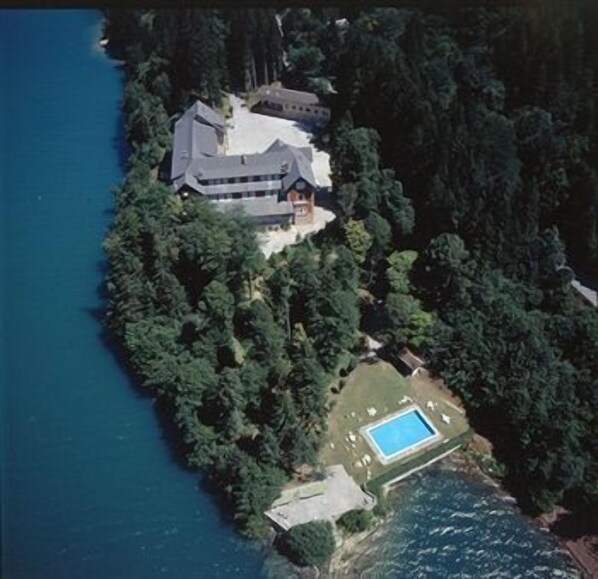 An aerial view of a large house surrounded by dense forest, with a swimming pool and a dock extending into the water.