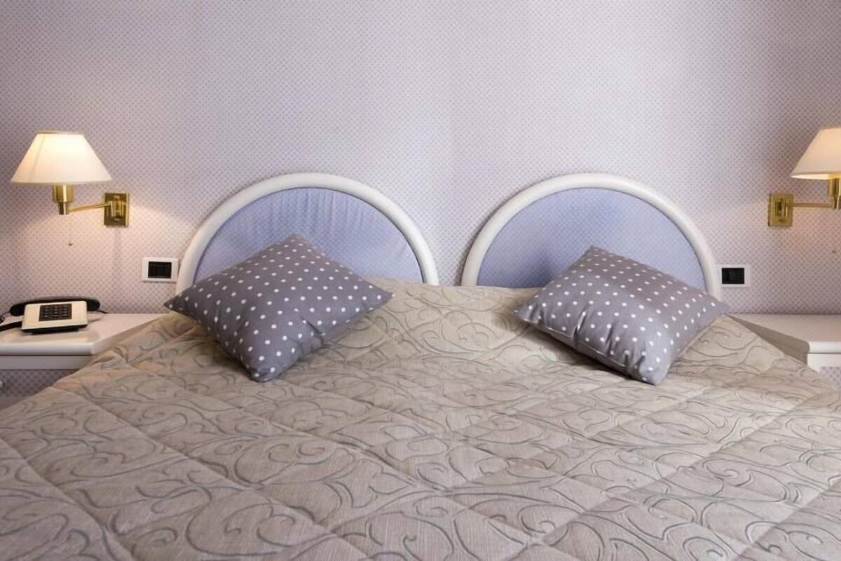 A double bed with a patterned bedspread, two pillows with a polka dot design, two bedside lamps with gold fixtures, and a telephone on the nightstand.