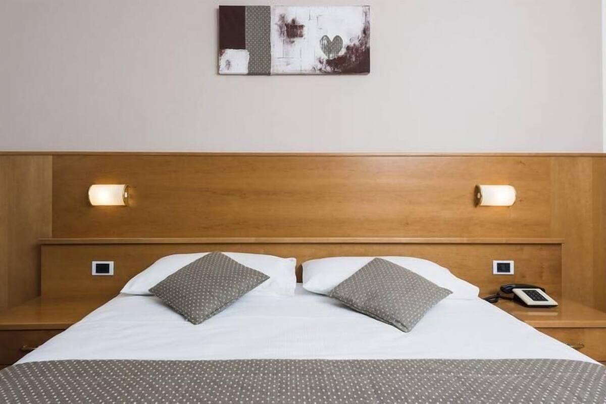 A hotel room with a wooden headboard, two bedside lamps, a bed with white bedding and gray pillows, and a small bedside table with a phone.