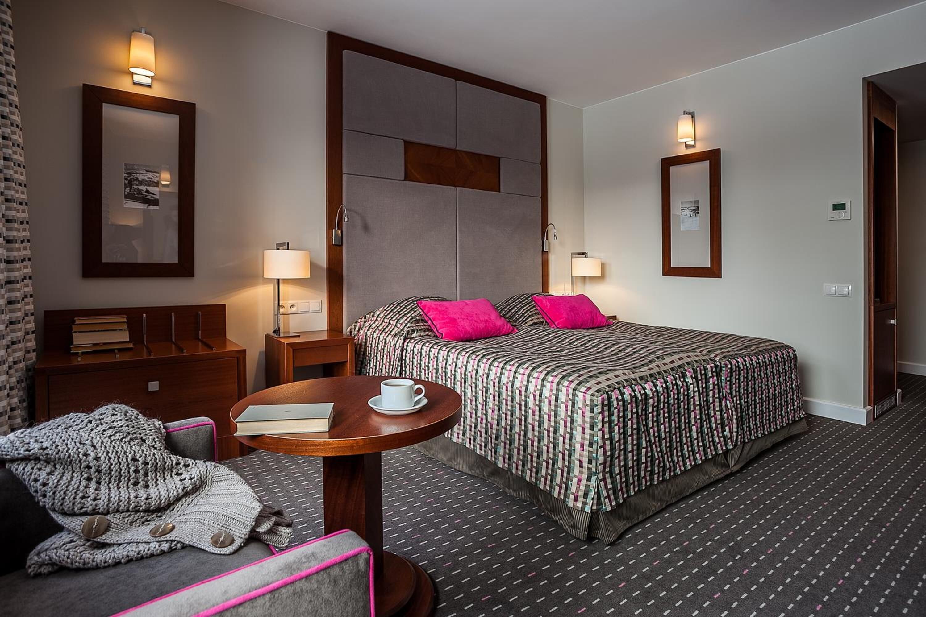 A hotel room with a bed, bedside tables, a desk, a chair, a round table with a cup, and a blanket.