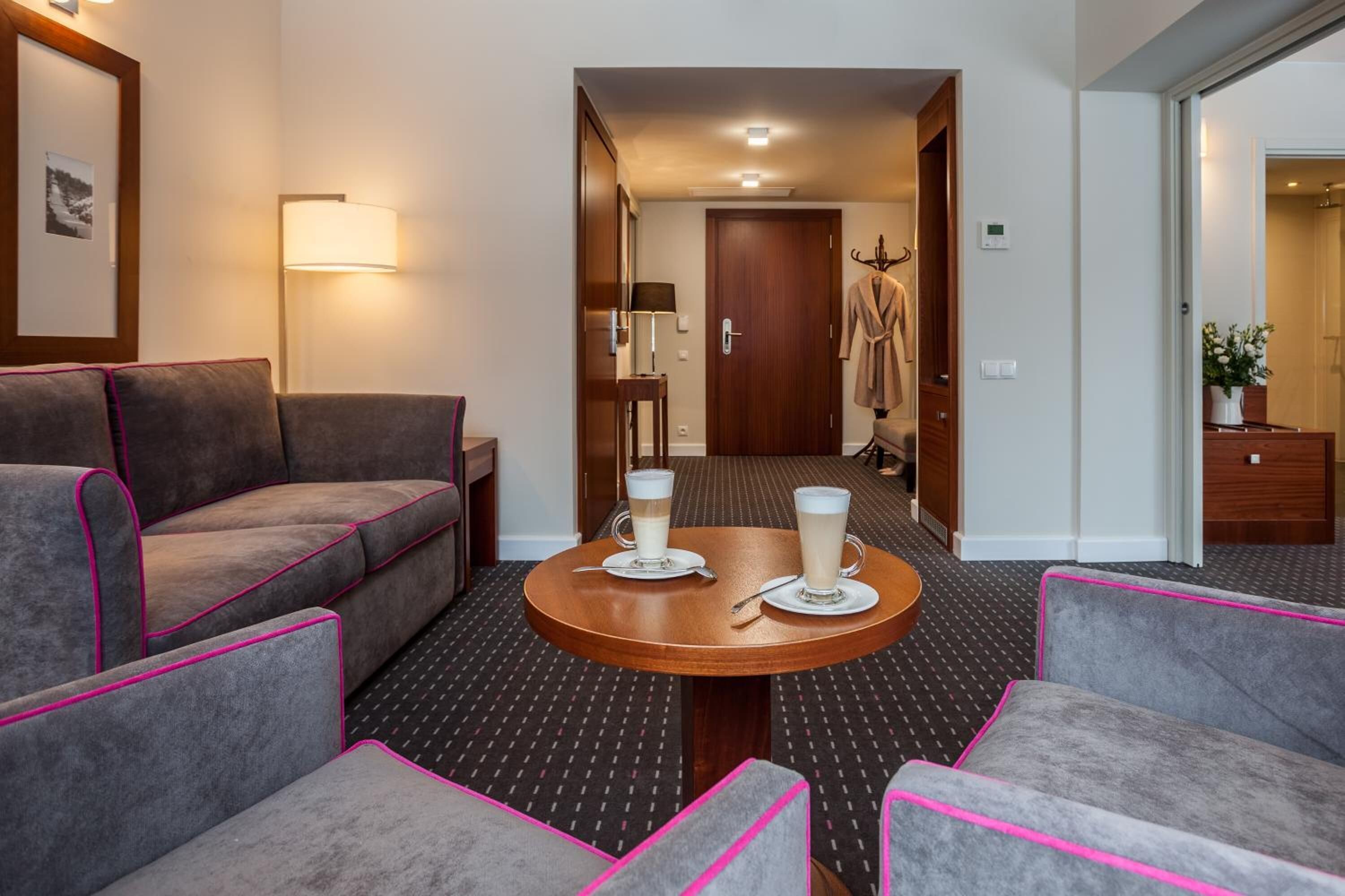 Une chambre d’hôtel avec un canapé, des fauteuils, une table ronde sur laquelle sont posées deux tasses de café, un peignoir accroché à la porte et une lampe fixée au mur.