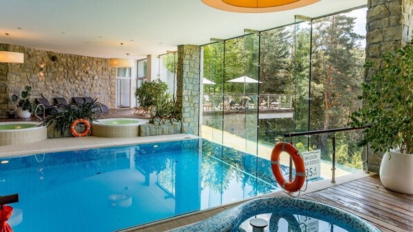 Una piscina interior con cerramiento de vidrio, pared de piedra y vista al bosque circundante.