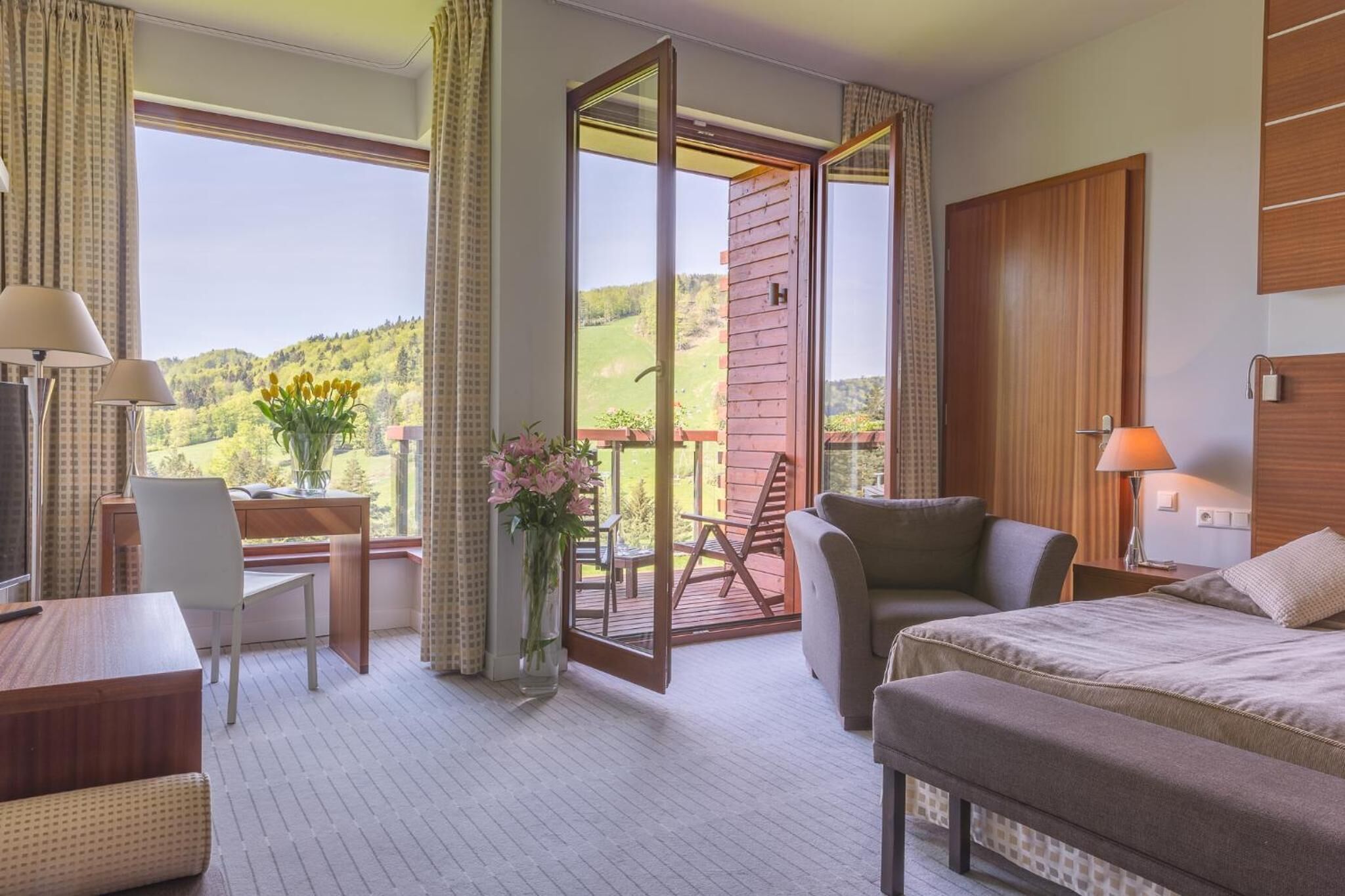 A hotel room with a bed, a desk, a chair, a sofa, a balcony with a view of greenery, and a window with curtains.
