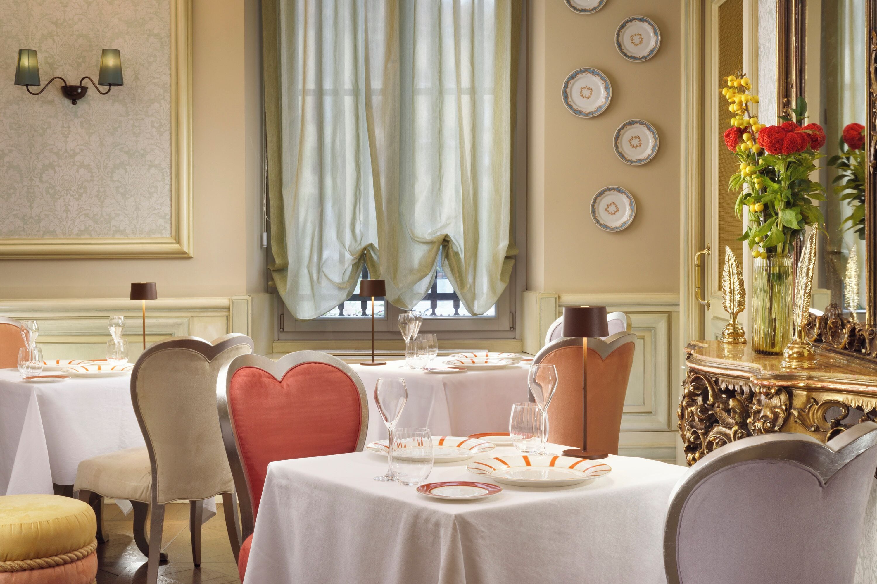 Un comedor con mesas dispuestas para el servicio, sillas, un ventanal con cortinas y un espejo decorativo con un florero.