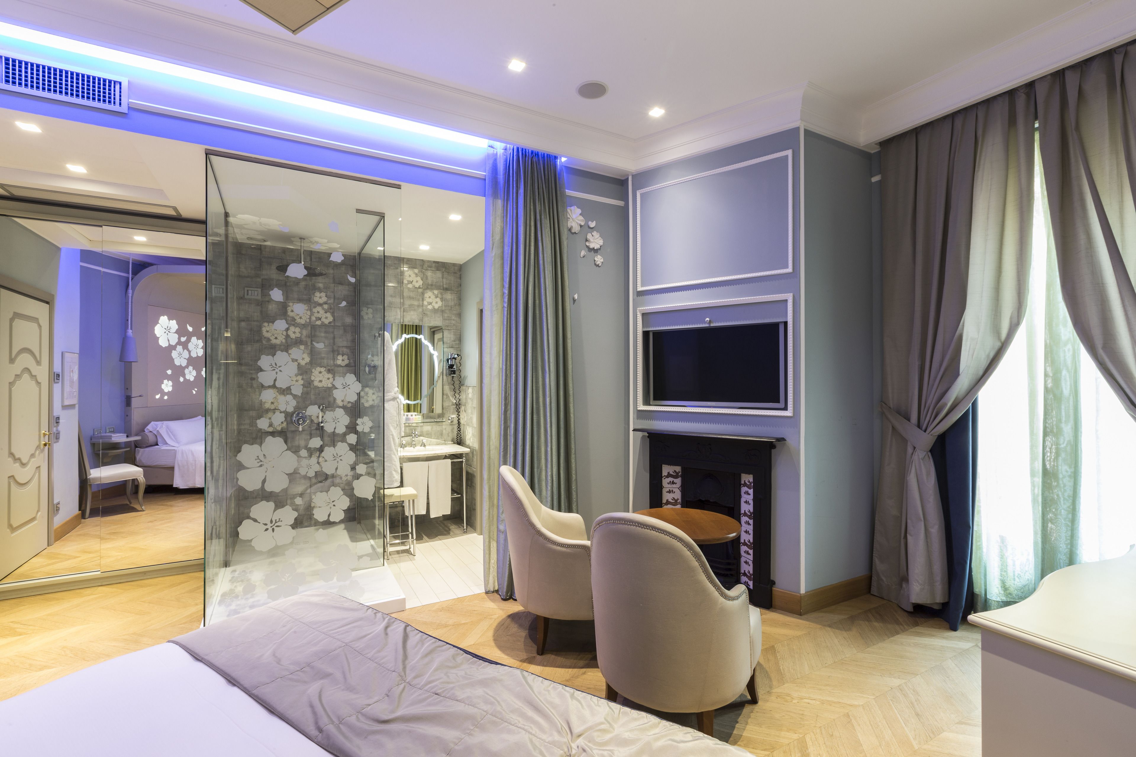 A modern hotel room with a bed, a seating area with two chairs, a TV mounted on the wall, and a fireplace.