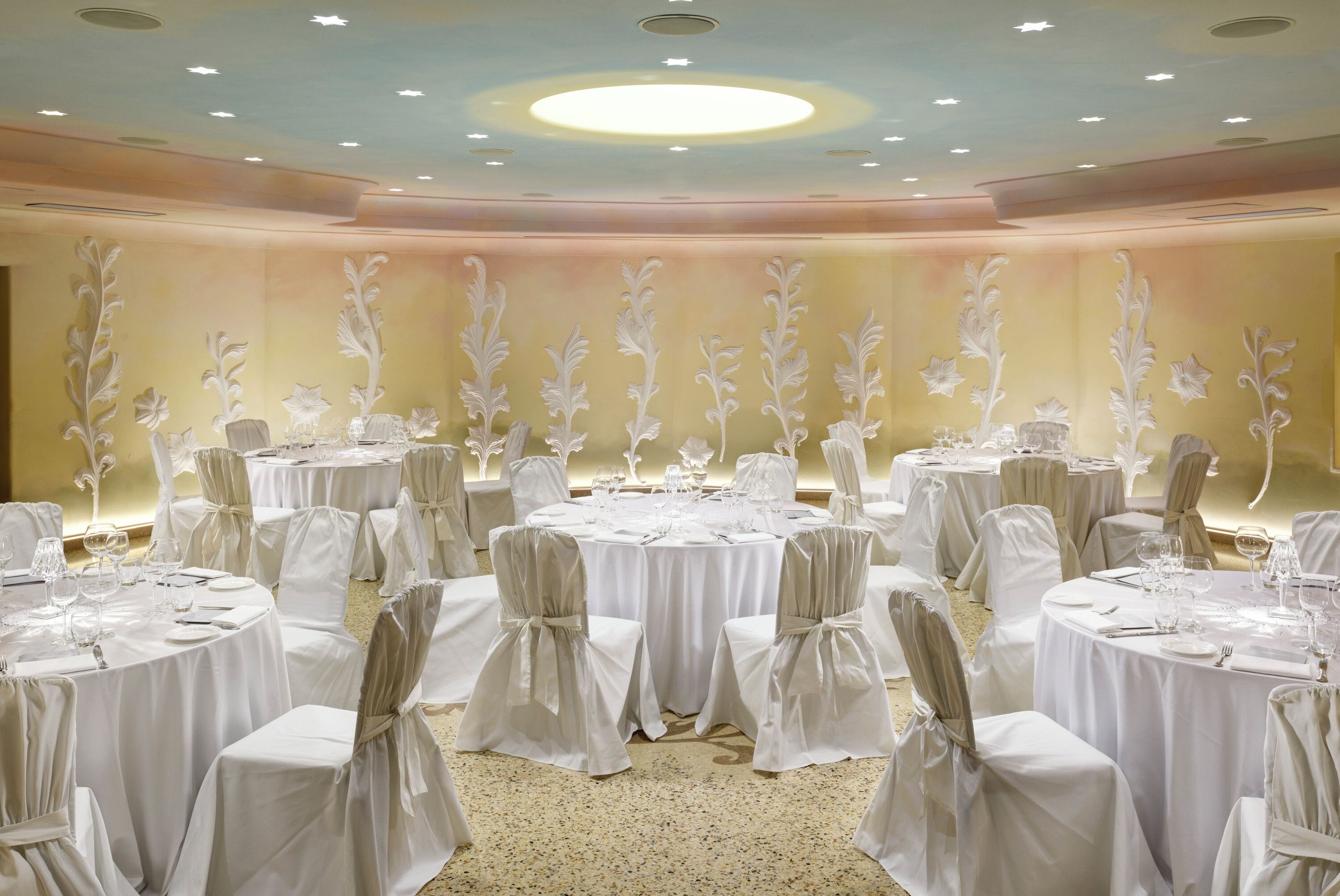Een feestzaal met ronde tafels, witte tafelkleden en stoelen met witte hoesjes.