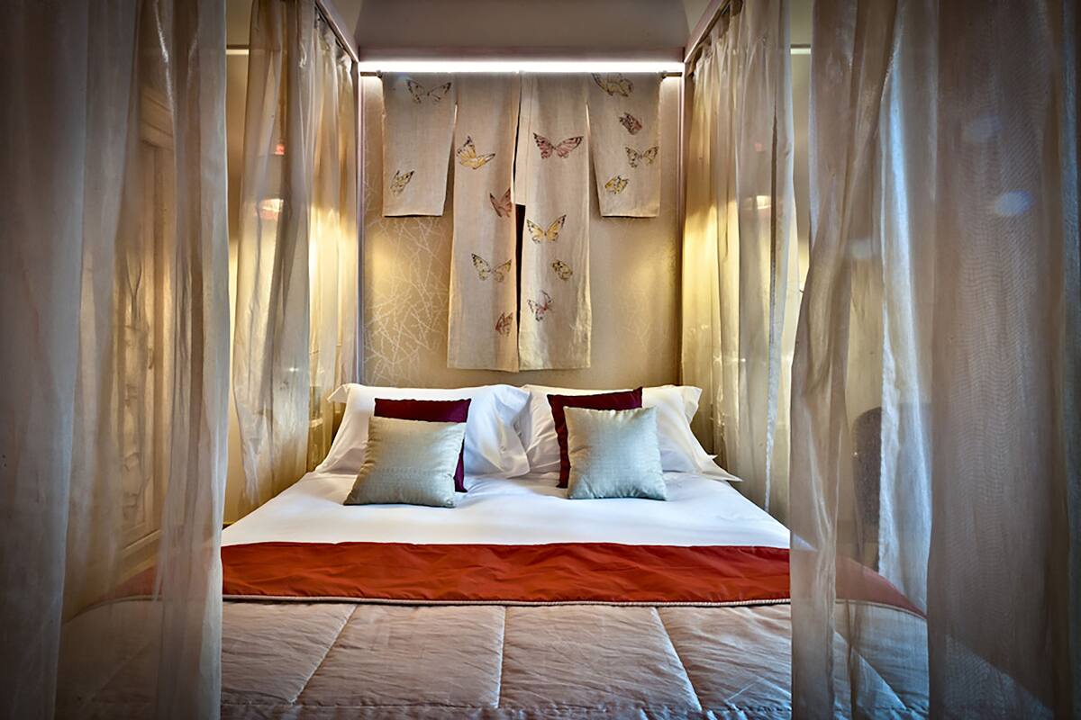 A neatly made bed with white linens and a red accent, surrounded by sheer curtains with butterfly patterns.