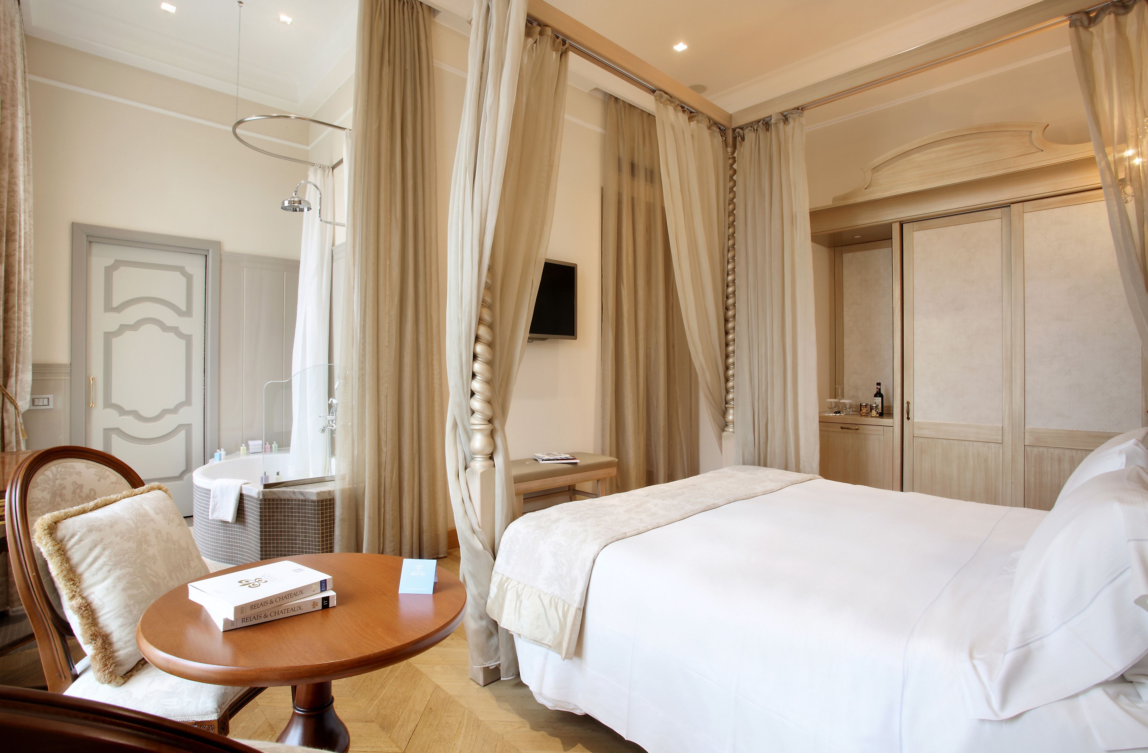 Une chambre d’hôtel comprenant un lit, une table de chevet, une chaise, une petite table avec un livre, une télévision et une salle de bain avec une baignoire et un miroir.