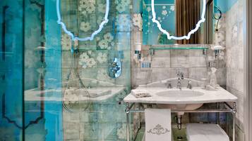 A bathroom with a glass shower enclosure, a white sink with a glass shelf, and a mirror with a floral design.