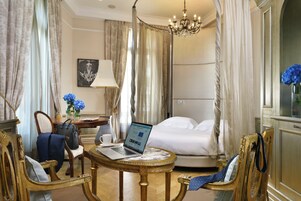 Een hotelkamer met een bed, een laptop op een ronde tafel, een stoel, een bureau met een lamp en een vaas met bloemen.