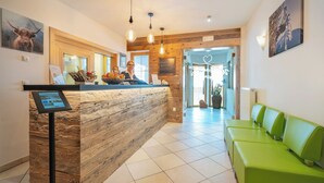 A reception area with a wooden reception desk, a digital information kiosk, a green seating area, and a view of the entrance. - Hotel Apartment Haus Gitschberg (Vandoies)