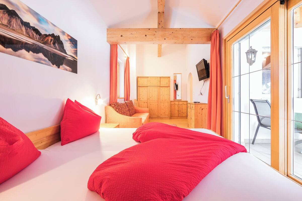 A bedroom with a bed, red pillows, a wooden headboard, a painting, a wardrobe, and a sliding glass door to a balcony.