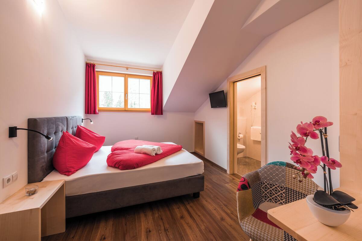 A modern bedroom with a bed, a bedside table, a window with curtains, a TV mounted on the wall, and a small table with a vase of flowers.
