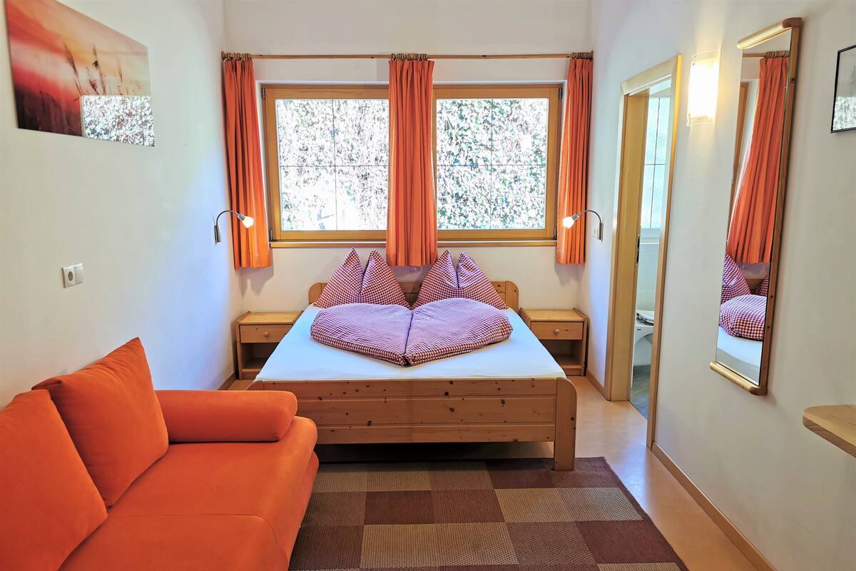 A bedroom with a bed, orange curtains, a mirror, a window, and a sofa.