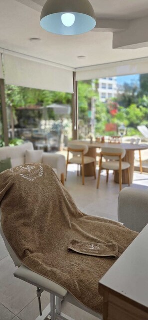 A modern room with a covered chair, a dining area, and a view of greenery and buildings outside. - Palmen Apart Hotel (Alanya)