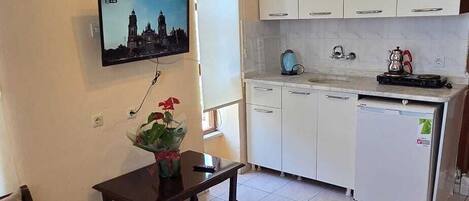 Una sala de estar con un sofá, una mesita con un florero, un televisor montado en la pared y una zona de cocina con armarios y un refrigerador.