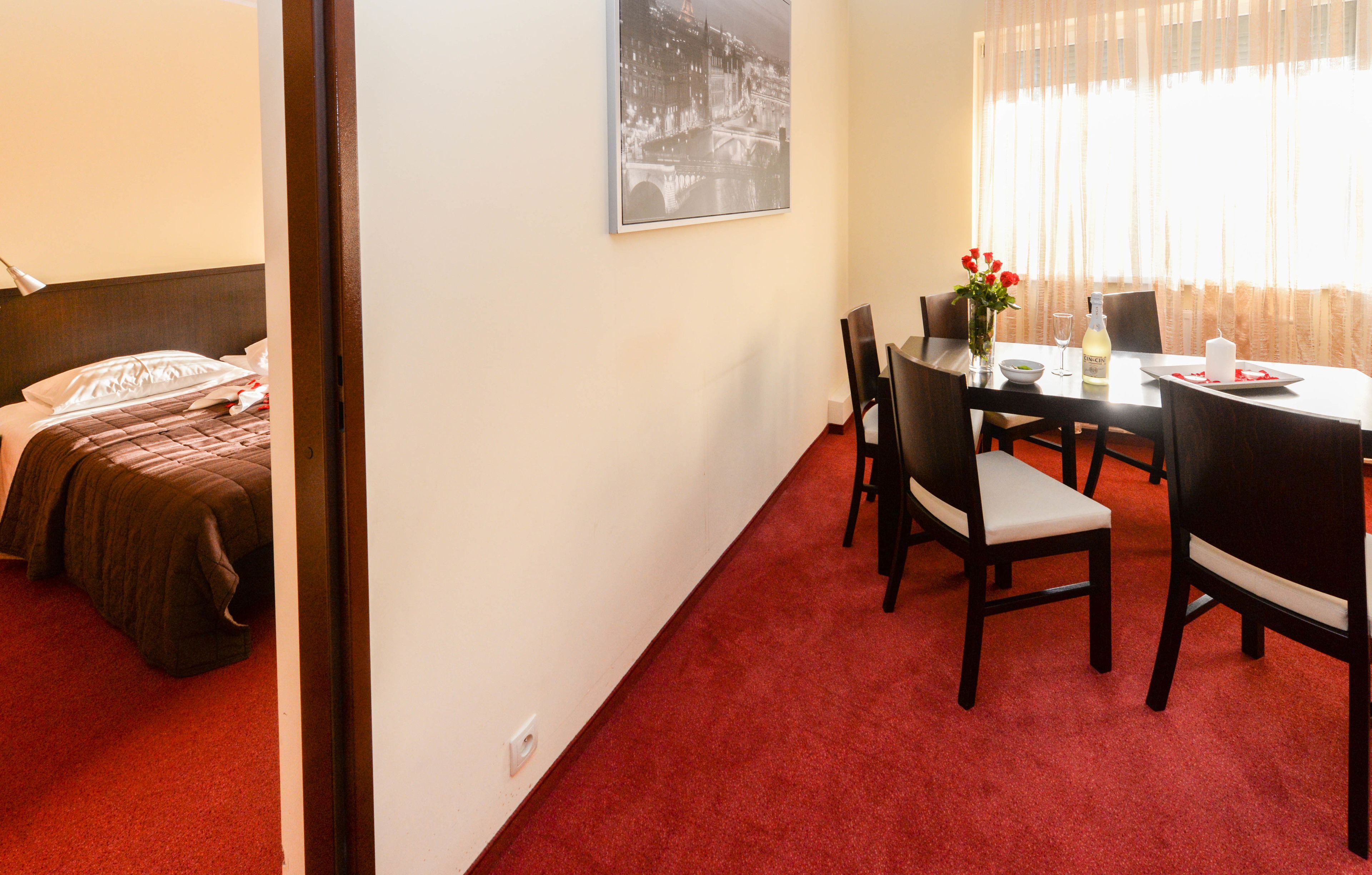 Una habitación de hotel con cama, una mesa de comedor con sillas, un florero con flores rojas, una botella de vino y un cuadro en la pared.