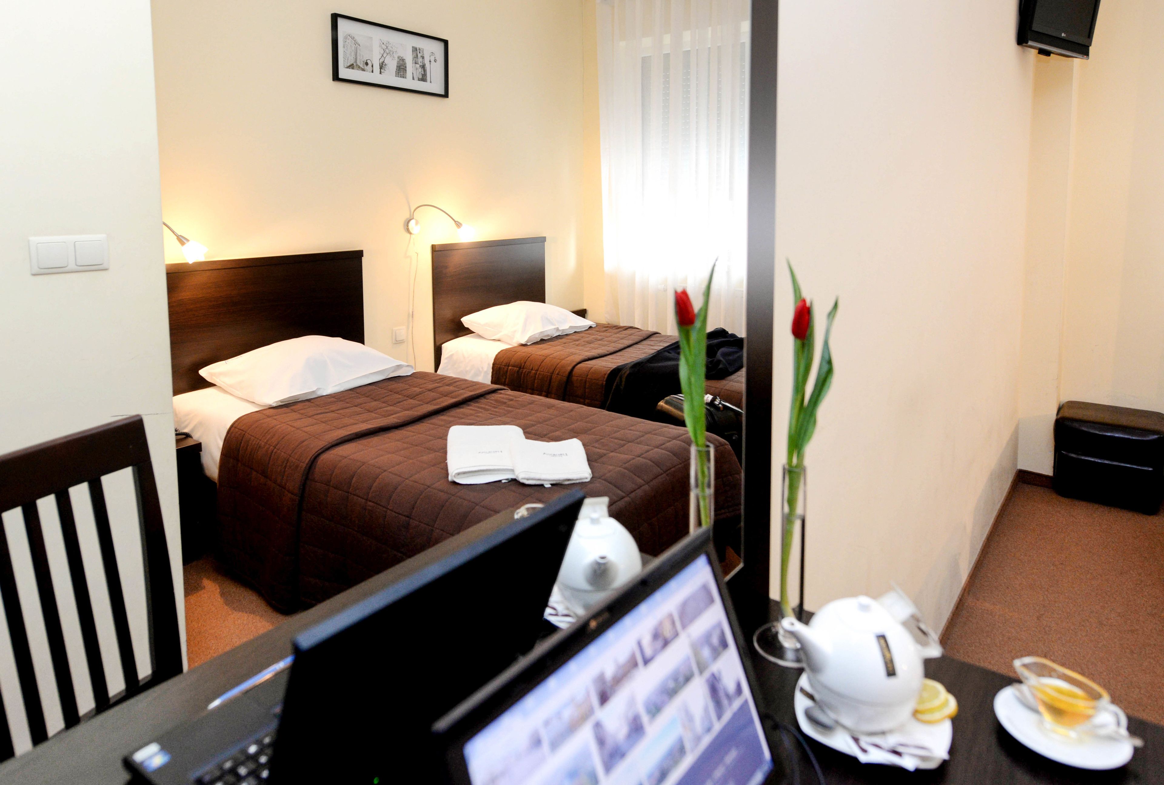 Ein Hotelzimmer mit zwei Betten, einem Schreibtisch mit Computer, einer Kaffeemaschine und einer Vase mit Blumen.