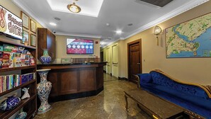 Un hall de hotel con mostrador de recepción, un sofá azul, un mapa en la pared y una estantería con libros y jarrones decorativos.