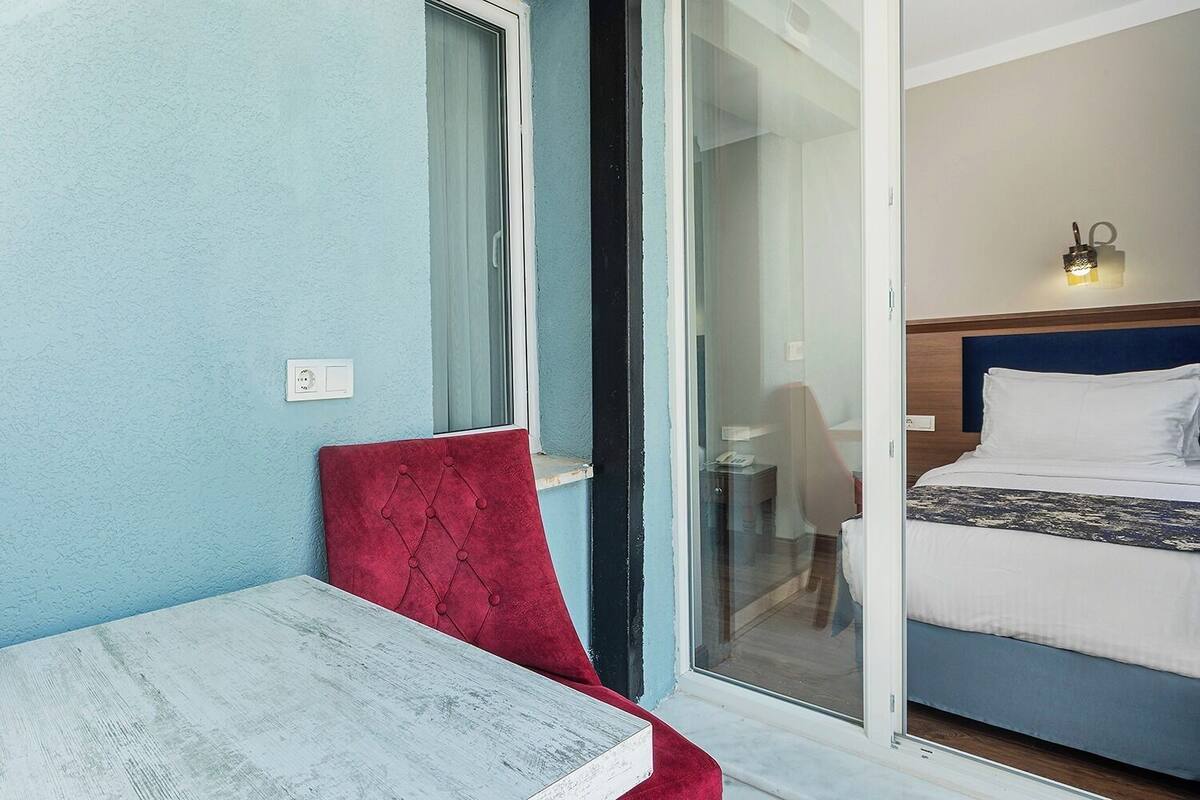 Une chambre d’hôtel avec un lit, une chaise rouge, une table et une porte-fenêtre coulissante donnant sur un balcon.