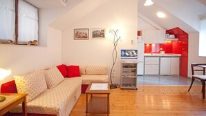 Uma sala de estar com sofá, uma mesa de madeira pequena, uma televisão e uma cozinha ao fundo.