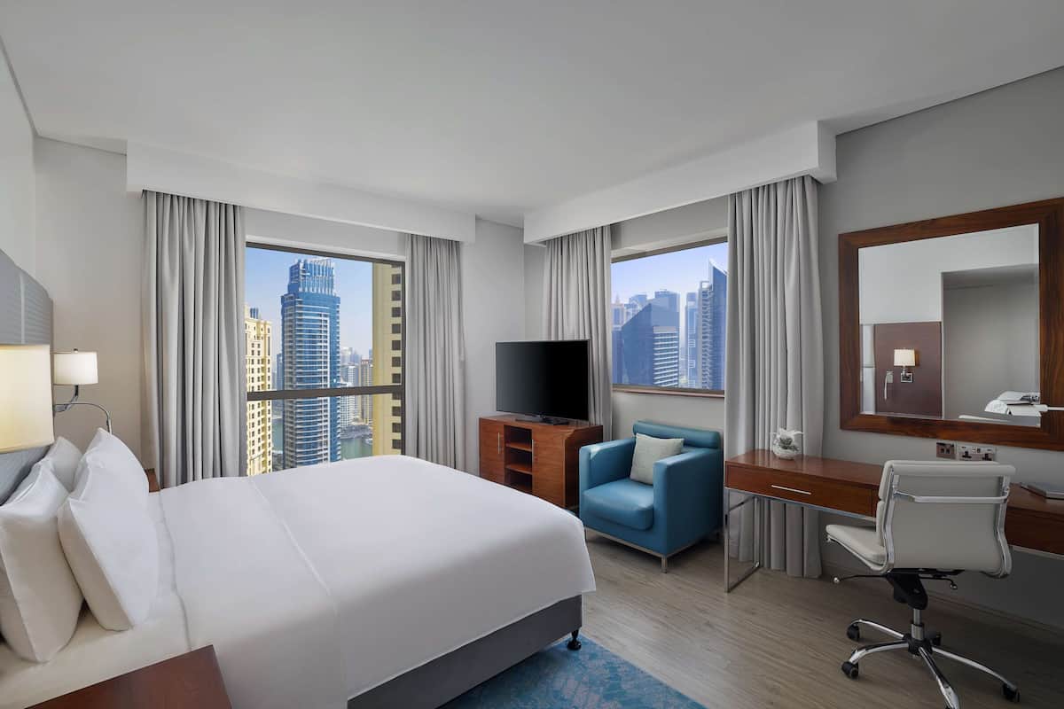 A hotel room with a large bed, a desk, a chair, a TV, and a city view.