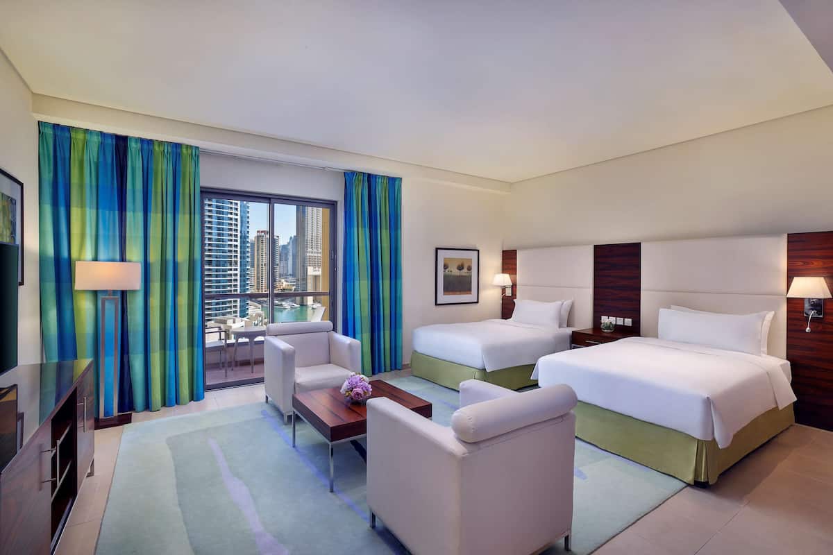 A hotel room with two beds, a desk, a chair, a TV, and a view of the city.