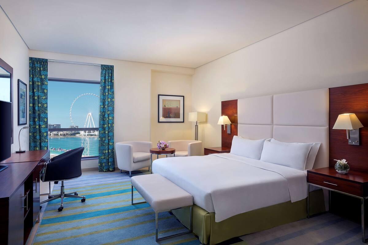 A hotel room with a large bed, a desk, a chair, a small table with a vase, and a view of a Ferris wheel.