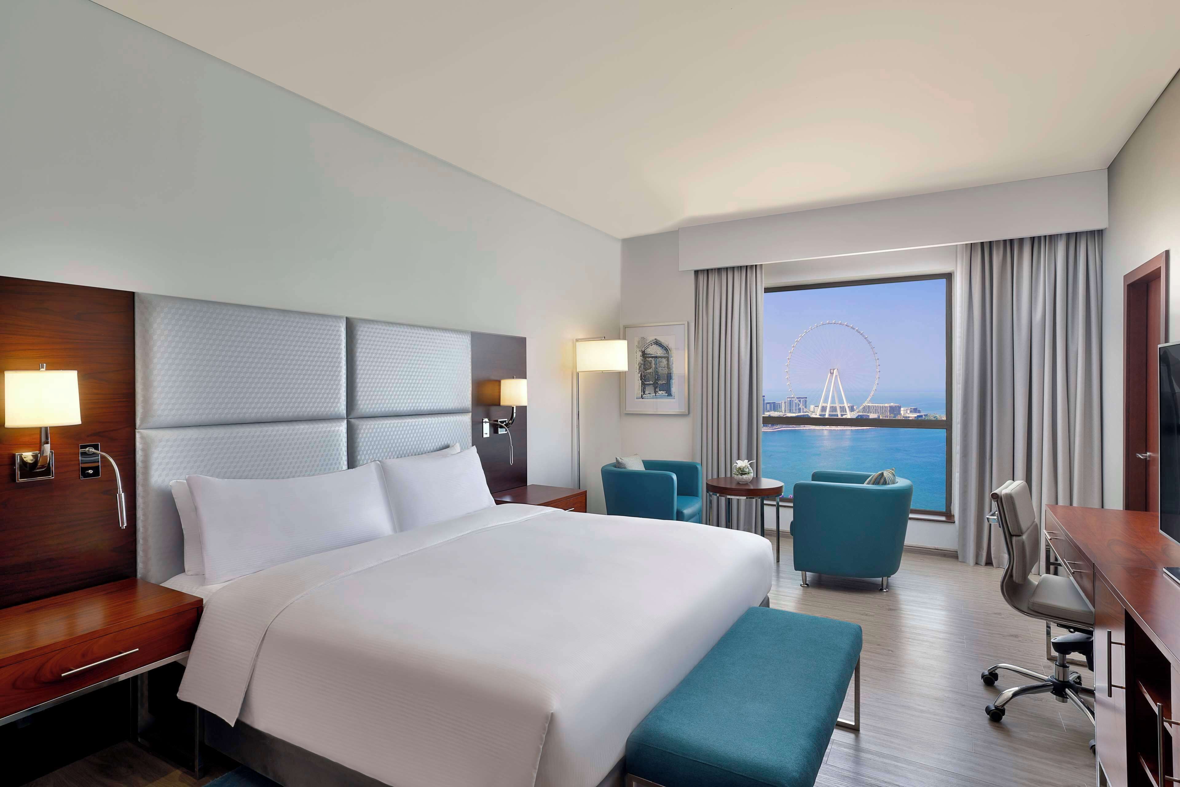 A hotel room with a large bed, a desk, a chair, a small table, and a view of a Ferris wheel and water.