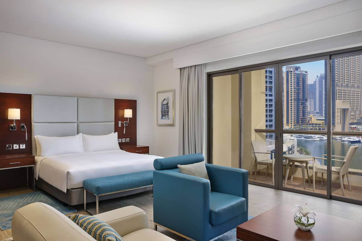 A hotel room with a large bed, a blue armchair, a small table with a vase, and a view of a cityscape through a large window.