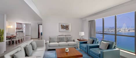 Una habitación de hotel moderna con un gran ventanal que ofrece vistas a una noria y al agua. Hay un sofá, un sillón azul y una mesa de centro de madera.
