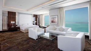 Una habitación de hotel moderna con un gran ventanal que ofrece vista al mar, una mesa de centro de vidrio, sofás blancos y una alfombra estampada.