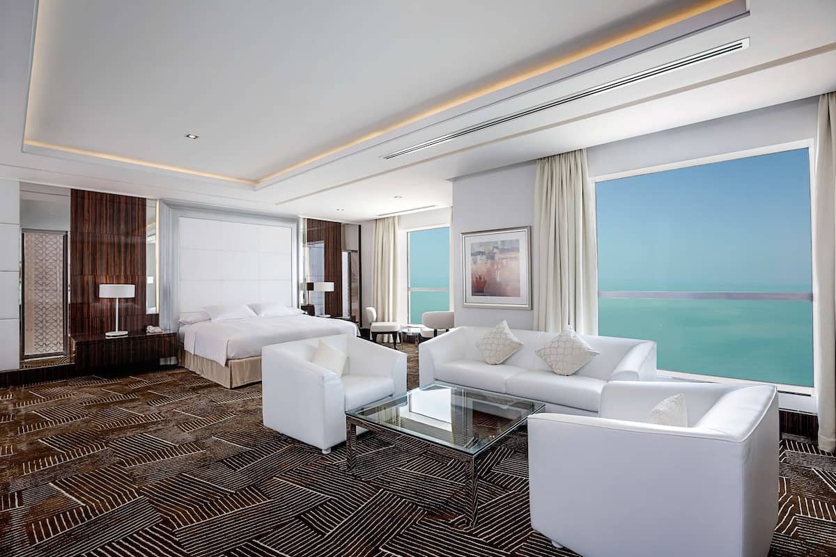 A modern hotel room with a large window offering a view of the sea, a glass coffee table, white sofas, and a patterned carpet.