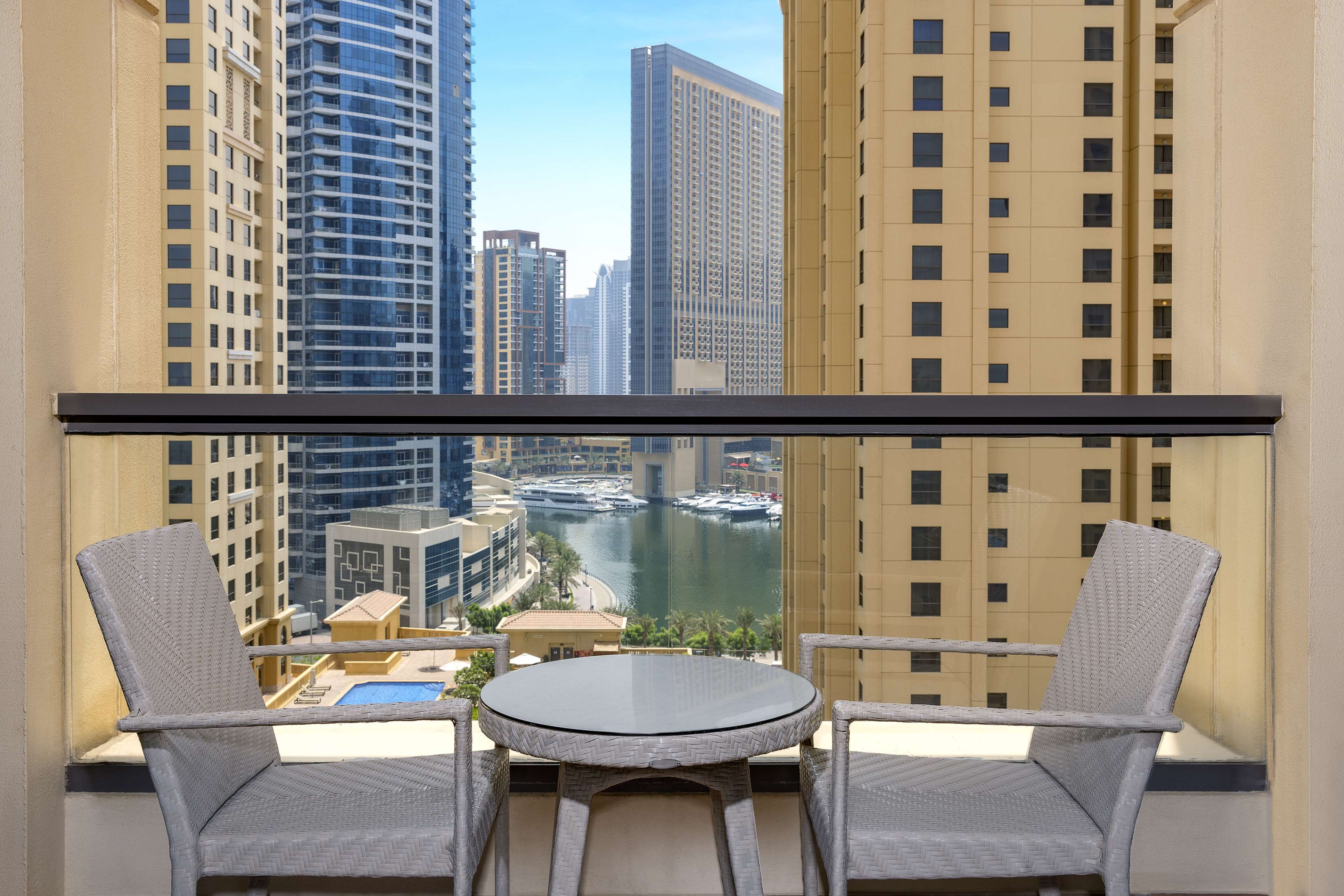 A balcony with two wicker chairs and a small table, overlooking a marina with boats and high-rise buildings.