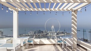 Una terraza en la azotea con mobiliario de mimbre blanco, guirnaldas de luces y vistas a una noria y al paisaje urbano.