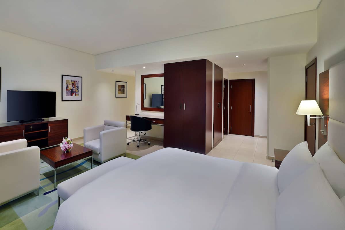 A hotel room with a large bed, a desk with a computer, a TV, and a chair. There are framed pictures on the wall and a lamp on the bedside table.
